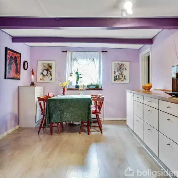 Et køkken-alrum med lilla vægge har et lille spisebord dækket med en grøn dug. Fire røde stole omkranser bordet. Til højre står en hvid køkkenenhed med en kaffemaskine ovenpå.