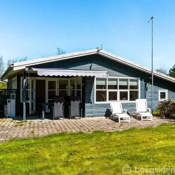 Et sommerhus med blå facade står på en velplejet græsplæne. Foran huset er der en terrasse med liggestole og spisebord. Omkring huset er der grønne buske og træer.