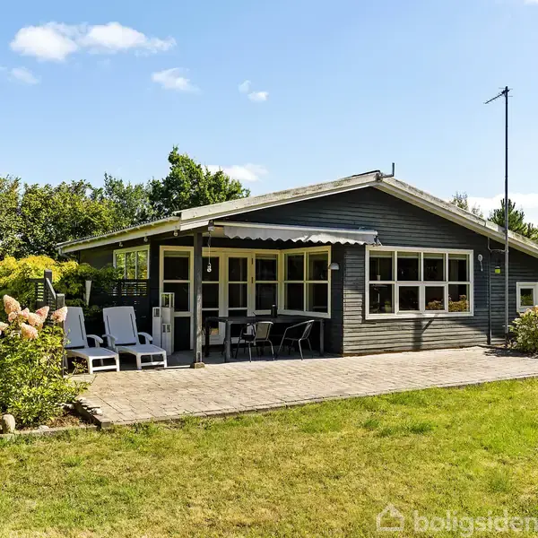 Et lille træsommerhus står stille på en græsplæne. Der er liggestole og blomsterbede på terrassen, omgivet af grønne buske og træer under en klar blå himmel.