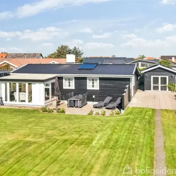 Et sort, enkeltplanshus med solceller på taget har en terrasse med havemøbler foran, beliggende i en pæn, grøn have med en bred græsplæne. Solrigt vejr.