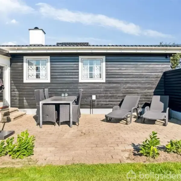 Terrasse med to liggestole ved siden af et bord og stole. Sorte træfacader omgiver terrassen med en lille græsplæne og planter i forgrunden. Klart vejr med lidt skyer.