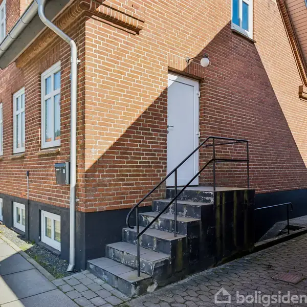 Rødt murstenshjørnehus med hvide vinduer har en metaltrappe, der fører til en hvid dør. Beliggende ved en fortovssti på en solrig dag.