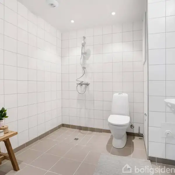 Toilet står ved siden af en bruser på en væg med hvide fliser. En håndvask er placeret til højre. En træbænk med en plante står til venstre.