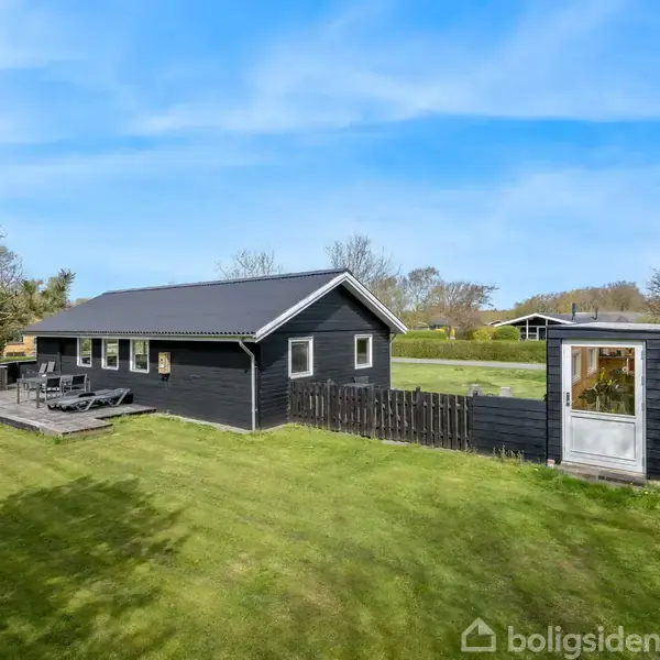 Et sort sommerhus med terrasse står i en grøn have omkranset af træer. Bord og stole er placeret på terrassen under en blå himmel.