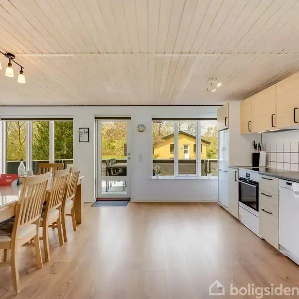 Lyst køkken-alrum med spisebord og stole til venstre. Køkkenet til højre har hvidevarer og lyse skabe. Store vinduer og en dør fører ud til en terrasse med udsigt til et gult hus.
