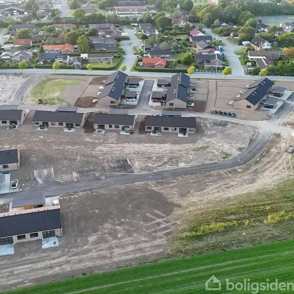 Rækkehuse under opførelse i et nybygget boligområde, hvor flere strukturer allerede står, omkranset af byggepladser og grønne marker. Boligkvarterer og veje kan ses i baggrunden.