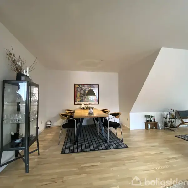 Spisebord omgivet af stole står på et tæppe i et minimalistisk rum med lyst trægulv. Glasskab til venstre indeholder glas. Væggene er lyse, og der hænger et billede.