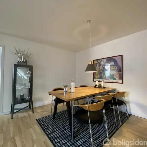 Et spisebord med fire stole står på et sort-hvidt tæppe i en spisestue. En lampe hænger over bordet, og et maleri pryder væggen. En glasvitrine står i hjørnet.
