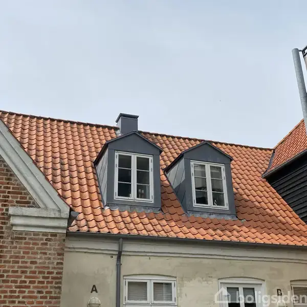 Røde teglsten på et hustag med to kvistvinduer. Bygningen er lavet af mursten og pudset væg. En altan ses til højre. Ingen tekst udover "Boligsiden."
