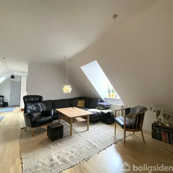 En stue med sort sofa og lænestol omkring et træbord, oplyst af en hængelampe. Skråvæg med vindue bagved, planter og pokaler i hjørnet. Trægulv med tæppe.