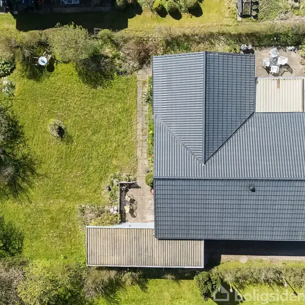 Hus med sort tag set oppefra, omgivet af frodig have med græsplæne og træer. Der ses en terrasse med havemøbler og en indkørsel med parkerede biler.