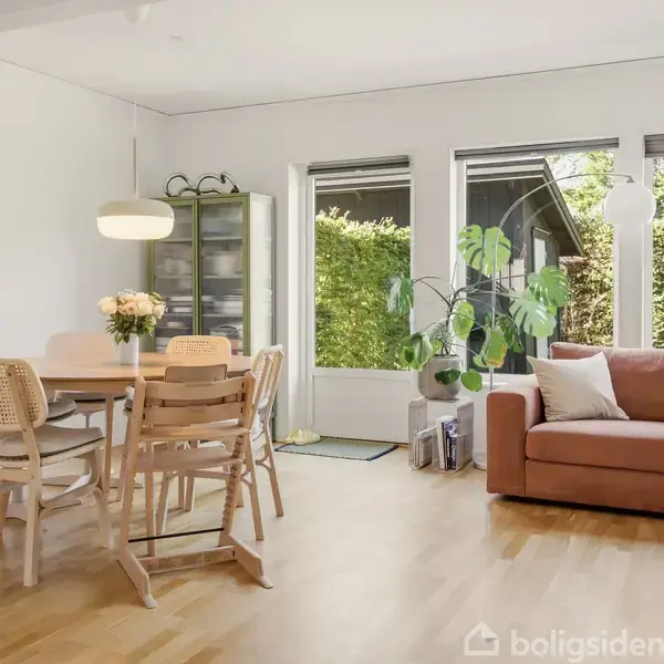 Sofagruppe og spisebord står i en lys stue med trægulv. Ét hjørne har tre store vinduer, der giver udsigt til en grøn have. En plante står ved sofaen.