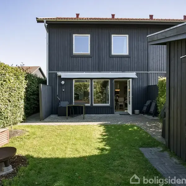 Toetages hus står i haven, hvor dets facade vender mod en græsplæne omkranset af hæk. En terrasse foran huset indeholder en stol og et bord.