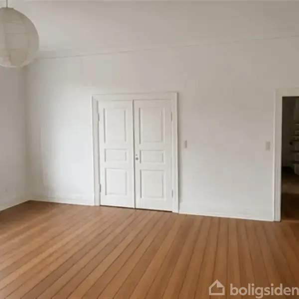 Et rummeligt værelse har et træplankegulv og hvide vægge. En stor, rund loftlampe hænger fra loftet. Der er en radiator under det store vindue.