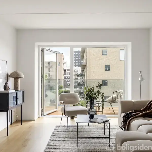 En stue med en beige sofa og sort sofabord vender mod en åben altandør. Store vinduer giver udsigt til moderne lejlighedsbygninger. En skænk står ved siden af.
