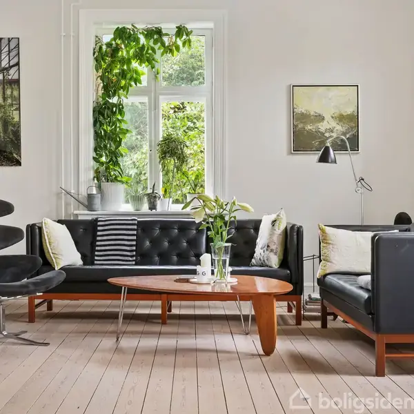 Stue med sorte lædersofaer og rundt træbord, omgivet af grønne planter og kunst på væggene. Lyst trægulv og et vindue giver rummet lys.