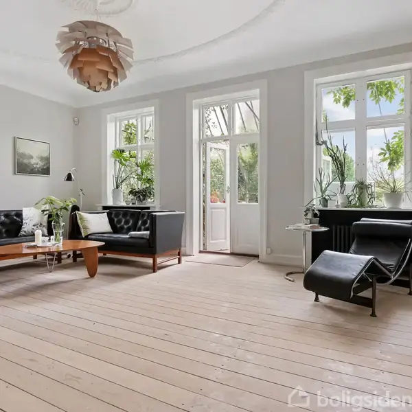 En stue med sorte lædersofaer står ved siden af et træbord. Store vinduer og en åben dør giver udsigt til træer udenfor. Planter dekorerer rummet.