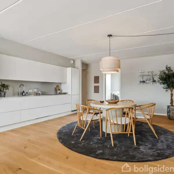 Spisebord med seks træstole står på et rundt gråt tæppe under en hængelampe. I baggrunden ses et moderne, hvidt køkken med indbygget ovn. En plante står til højre.