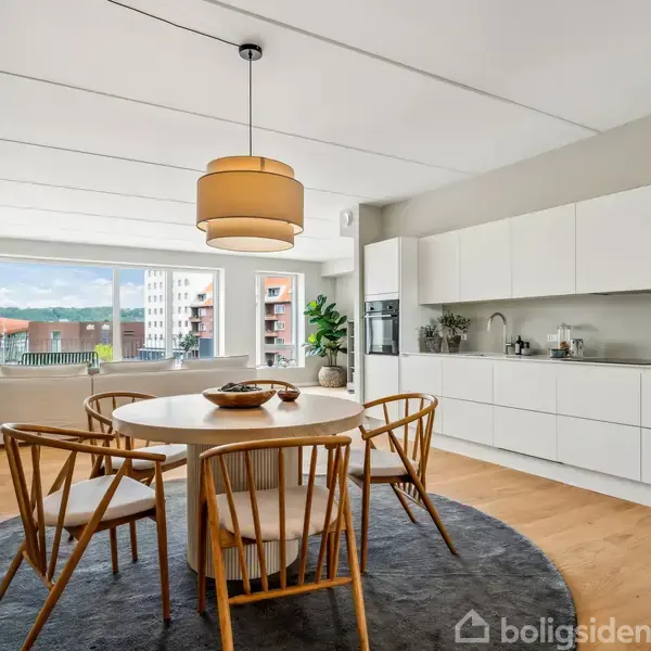 Rundt spisebord med fire stole står på et gråt tæppe. Køkken med hvide skabe er i baggrunden. Store vinduer viser en udsigt over en by med blå himmel.