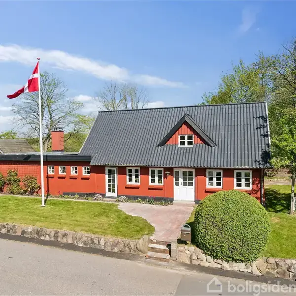 Et rødt hus står omgivet af en velholdt have med en dansk flagstang foran. Omkringliggende grønne træer skaber en fredelig atmosfære under en blå himmel.