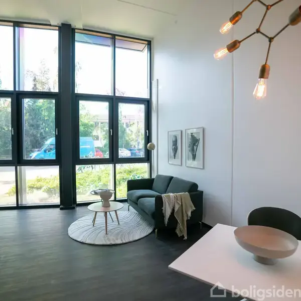 En moderne stue med en sort sofa ved et rundt tæppe, et skrivebord i hjørnet, store vinduer bagved, og en stilfuld lysekrone i loftet.
