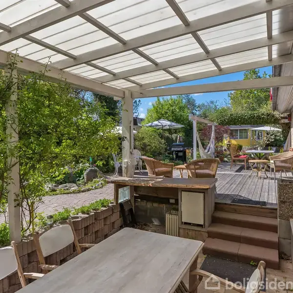 En overdækket terrasse med et bord og stole. Sollys stråler gennem taget. I baggrunden er der en hævet træterrasse med møbler, omgivet af frodig have og blå himmel.