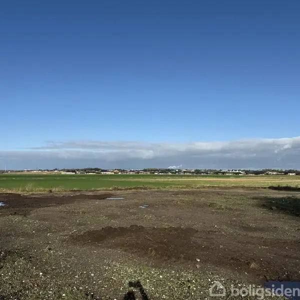 Feltet ligger ubevokset og tørt, spredte vandpytter ses på jordoverfladen. I baggrunden anes en lav bebyggelse under en klar, blå himmel med enkelte skyer nær horisonten.