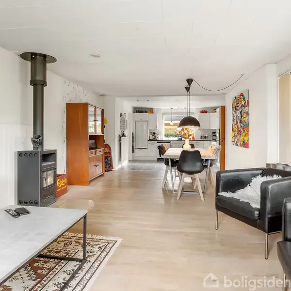 En åben stue har en sort brændeovn ved væggen. Midt i rummet står et spisebord med stole. Store vinduer på højre side giver udsigt til en have.