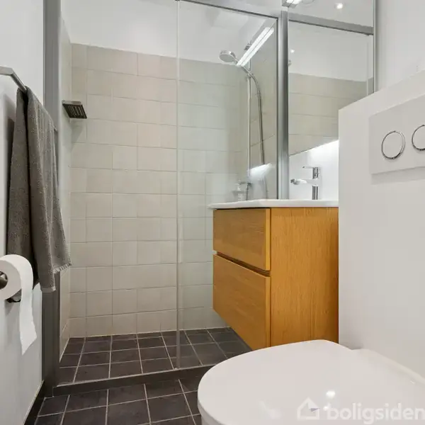 Toilet med to skylleknapper er i fokus. Dusjkabine bagved med glasdør og moderne indretning. Gråt håndklæde hænger ved siden af toiletrulleholder i minimalistisk badeværelse.