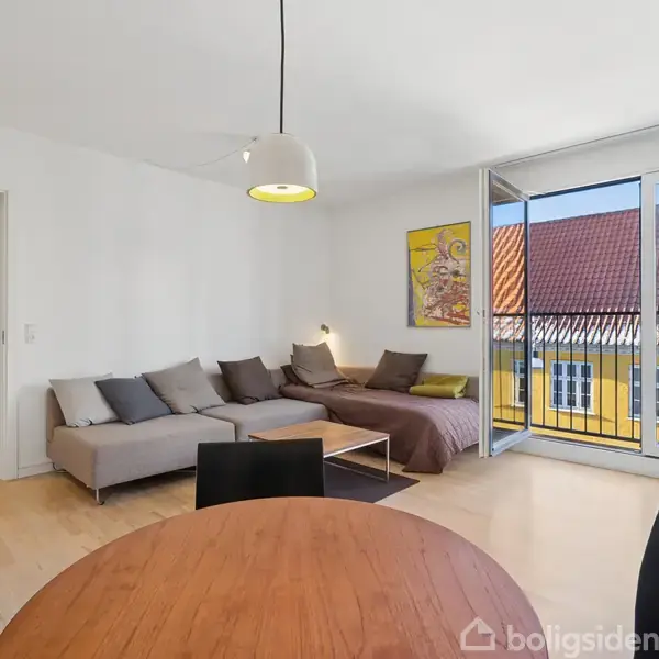 En stue med en grå hjørnesofa og sofabord. Et rundt spisebord står i forgrunden. Store vinduer åbner ud mod en altan med udsigt til et gult hus.