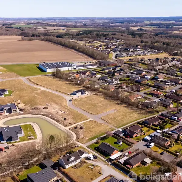 Bygninger set fra luften, ligger ved siden af en sø og åbne marker. Naboområdet består af en tætliggende boligudvikling med flere gader og huse. Landligt landskab i baggrunden.