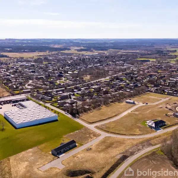 Byområde set fra oven med spredte bygninger og veje. En stor, rektangulær bygning befinder sig i forgrunden. Landskab fyldt med marker og skov i baggrunden under en klar himmel.