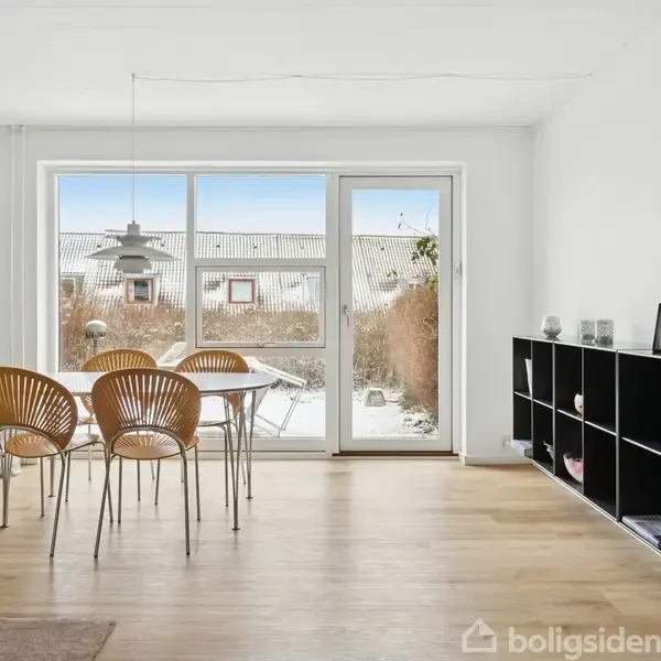 Spisebord med fire stole står ved et stort glasvindue med udsigt til en snedækket terrasse. På højre side ses en sort reol med flere rum.