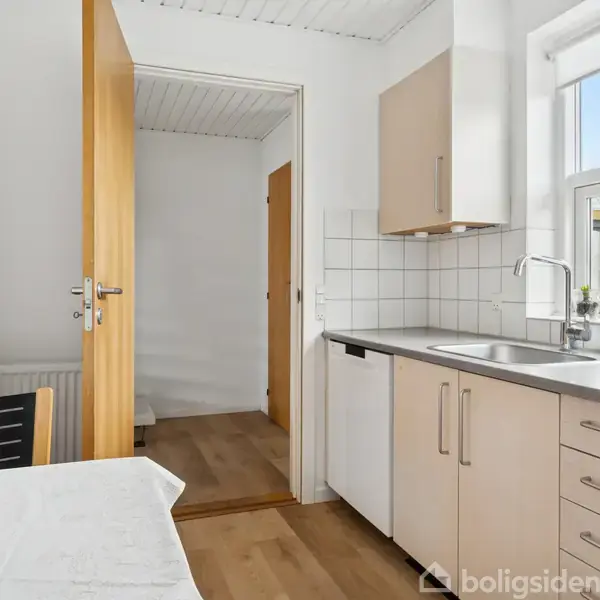 Et hvidt køkkenbord med vask ved et stort vindue; en åben dør fører til en gang. Et bord og stol står til venstre. Trægulv og hvidmalede vægge.