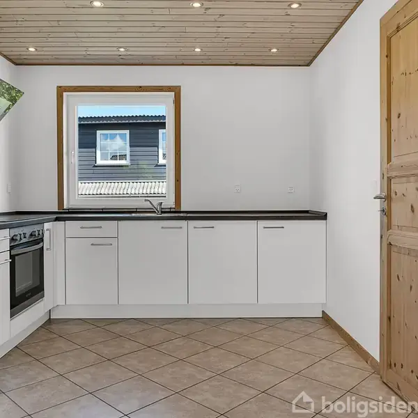 Køkken med hvide skabe og indbygget ovn. Vindue over vasken giver udsigt til et hus. Trægulv og -dør i traditionelt design. Loftspots giver belysning. Ingen tekst i billedet.