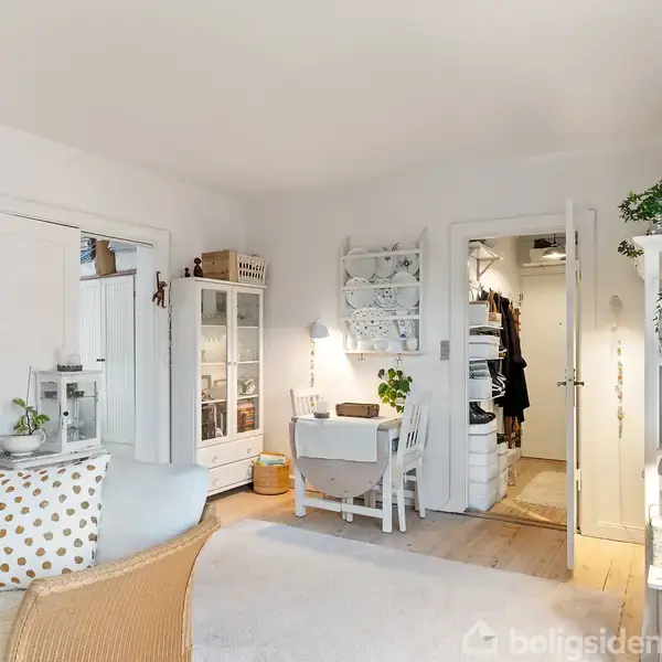 Stue med hvide vægge og trægulv indrettet med lys sofa og polstrede stole. Et skrivebord med stol er placeret ved væggen. Der er åbent skab og hylder med dekorationer.