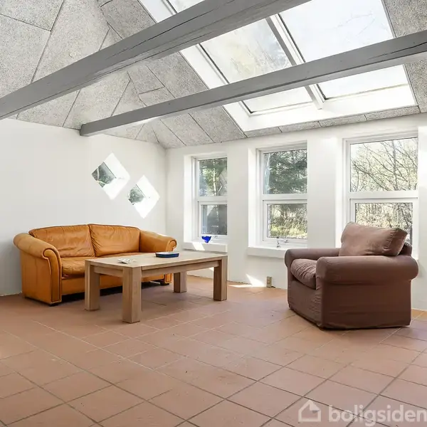 En stue med en orange sofa og en brun lænestol omkring et træbord. Der er lyse vægge, store vinduer og et skråt loft med ovenlys. En farverig maleri står ved siden af sofaen.