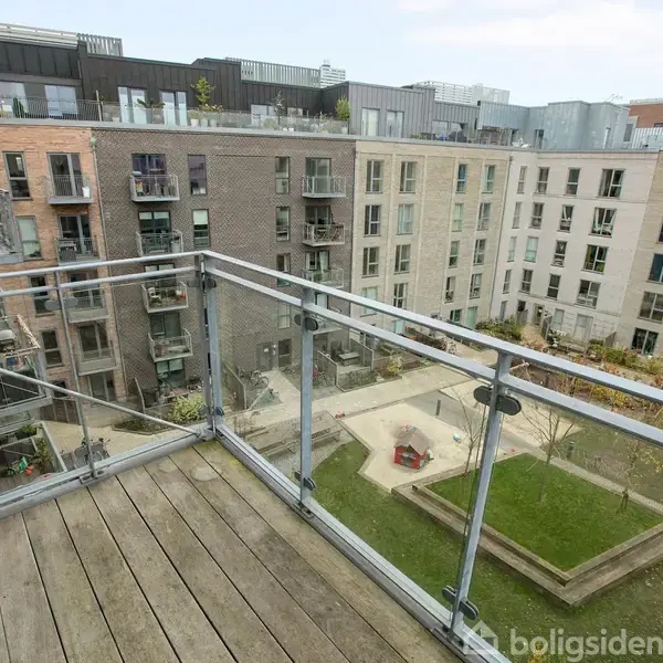 En balkon med glasrækværk tilbyder udsigt over en lukket gård med græsplæner, stier og beboelsesejendomme i baggrunden.