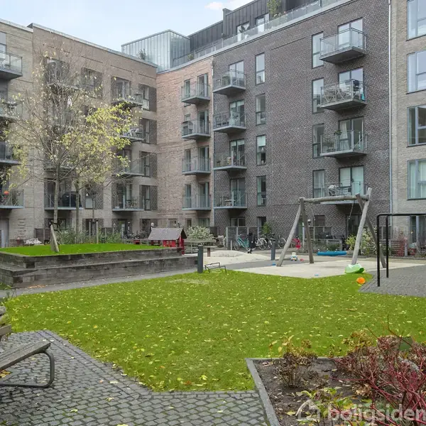 Bygning med altaner omkranser en indre gård med græsplæne og legepladsudstyr. En bænk står i forgrunden, og omgivelserne inkluderer træer og buske.