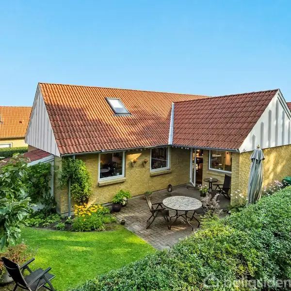 Et gult murstenshus med rødt tegltag står omgivet af en velplejet have. En terrasse med havemøbler ses foran huset, indrammet af veltrimmet hæk og grønne planter under en klar blå himmel.