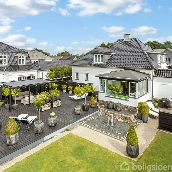 En villa med sort tag har en stor terrasse med planter og loungemøbler. Terrassen er omgivet af velplejede haver. I baggrunden ses flere huse under en skyet blå himmel.