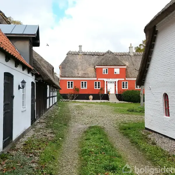 En stor, rød bindingsværksbygning med stråtag står foran. Den omkranses af to hvide sidebygninger med mørke tage. Vejen mellem dem fører ind til hovedhusets indgang.