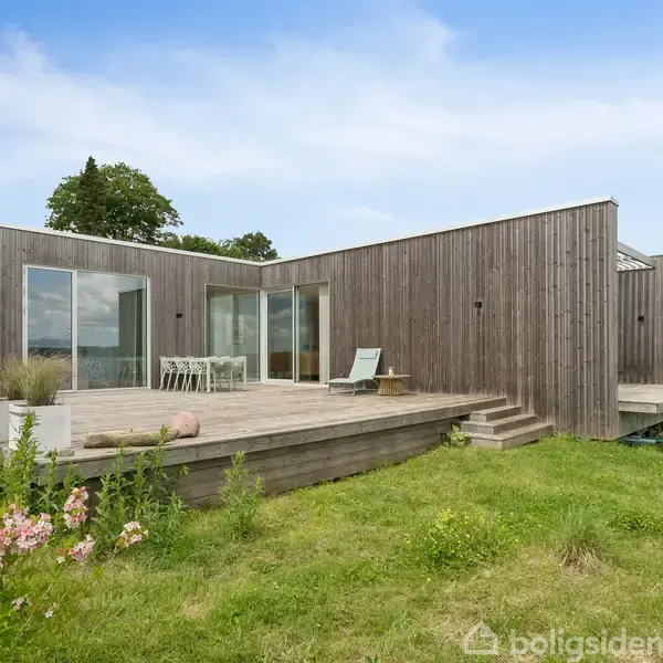 Træhus med store vinduer står stille på en hævet træterrasse. Terrassebord og stol ses. Omgivet af grønt græs og blomster med himmel og træer i baggrunden.