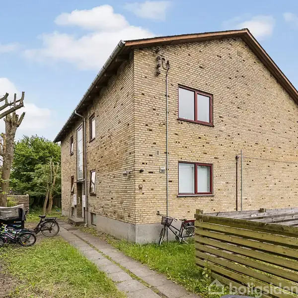 Toetagers murstenshus står i en græsplæne, omkranset af hegn. Cykler står parkeret ved siden af huset. Det er en klar dag med blå himmel og lette skyer.