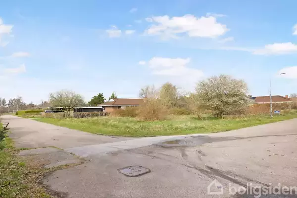 Vejen slynger sig gennem et beboelsesområde med græs og buske, omgivet af lave, røde murstenshuse under en blå himmel med få skyer.