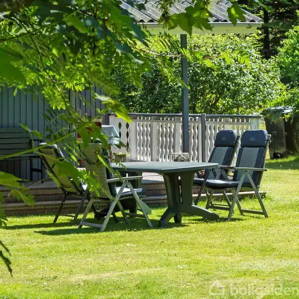 Et bord med fire stole står på en græsplæne i en have. Ved siden af er der et træhus med en terrasse og grønne buske i baggrunden.