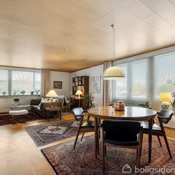 I et lyst stueområde står et rundt spisebord med fire stole. Rumindretningen inkluderer også lænestol, sofa og flere gulvlamper. Store vinduer giver udsigt til en have.