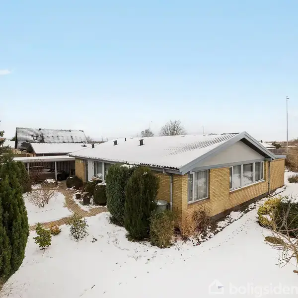 Et murstenshus dækket af sne står i en have med buske og træer. Himlen er klar blå, og der ligger sne på taget og jorden.