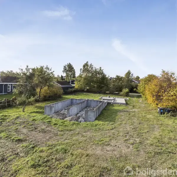 En ufuldendt byggeudgravning ligger stille i et grønt område med græs og spredt bevoksning. Omkring er der enkelte huse og træer under en klar blå himmel.