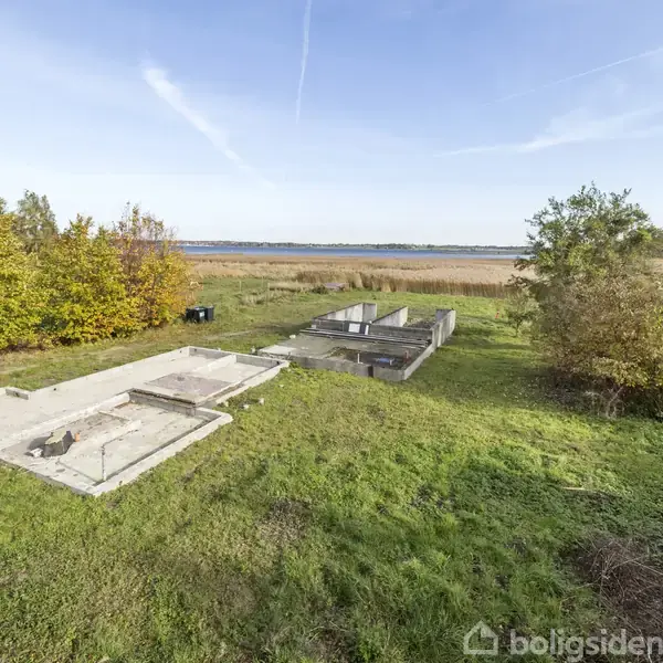 Betonfundament ligger stille på en græsplæne, omgivet af træer og buske. I baggrunden ses en sø med siv samt en åben, blå himmel.